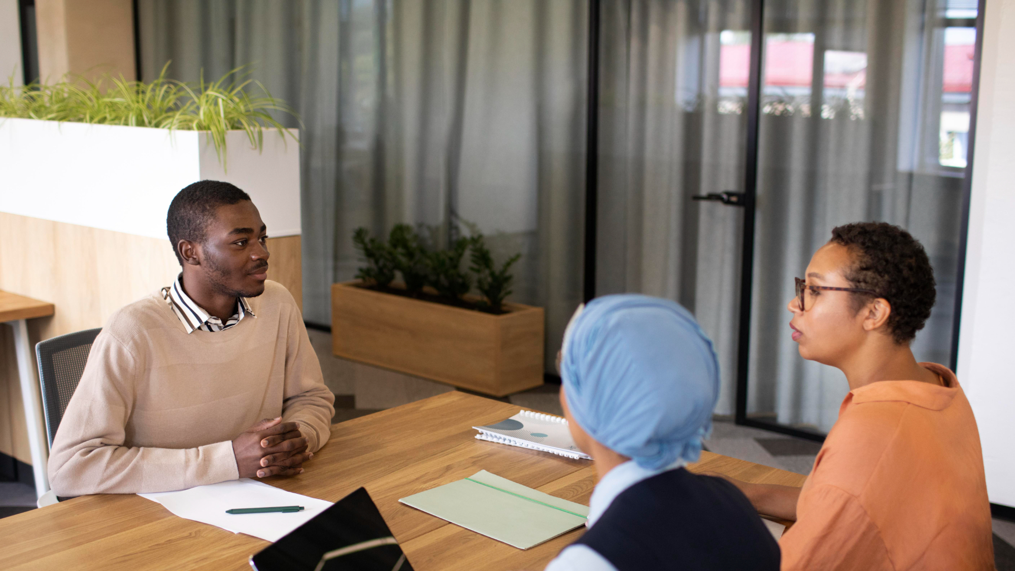 Three people in conversation across a table in a bright meeting room, one listening while another explains
